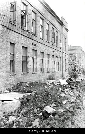 Photo d'archive vintage en noir et blanc des années 1950 ou 1960 montrant la façade extérieure en briques d'un bâtiment industriel à l'usine d'armature et d'isolation de Slavyansk. Le long atelier de l'usine dispose de plusieurs fenêtres en bois ouvertes le long du mur. Au premier plan, des tas de terre excavée et de gravats de construction indiquent un développement ou des réparations continus du territoire. Un aperçu historique authentique de l'infrastructure manufacturière soviétique, de l'architecture d'entreprise et de l'environnement industriel quotidien dans le Donbass paisible. Banque D'Images