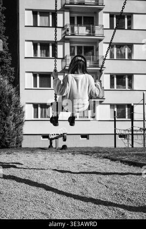 Photo en noir et blanc de l'enfant assis sur la balançoire dans l'aire de jeux urbaine avec bâtiment résidentiel en arrière-plan. Scène de style de vie quotidienne capturant l'enfant Banque D'Images