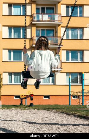 Enfant heureux assis sur la balançoire dans l'aire de jeux urbaine avec immeuble résidentiel en arrière-plan. Scène de style de vie quotidienne capturant l'enfance, le jeu Banque D'Images