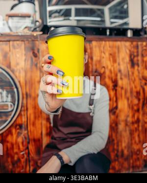 Un barista tient une tasse de café jaune dans un café. Le barista est une jeune femme aux cheveux courts, portant un pull gris et un tablier brun, assise devant Banque D'Images
