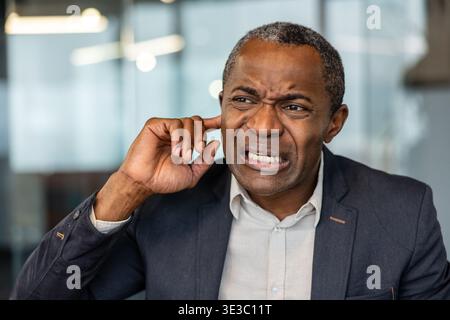 Homme mûr grimaçant, insérant un doigt dans son oreille, indiquant un bruit irritant, un bruit fort soudain, ou éprouvant des maux d'oreille, des acouphènes ou des problèmes d'audition au travail Banque D'Images
