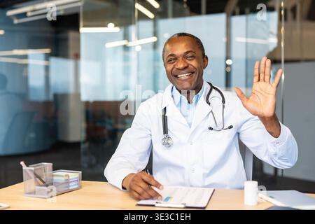 Médecin afro-américain portant une blouse de laboratoire blanche et un stéthoscope, souriant chaleureusement et agitant la main tout en étant assis à un bureau dans un cabinet médical moderne, fournissant des soins virtuels Banque D'Images
