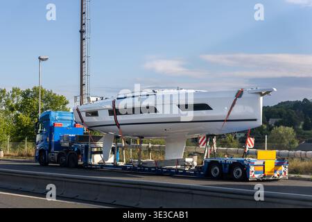 Camion lourd spécial transporte la coque de yacht à voile sur remorque basse le long de l'autoroute à travers la campagne. La coque sécurisée du yacht se déplace comme un convoi avec escorte Banque D'Images