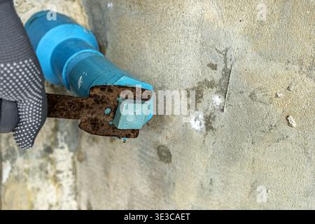 Gros plan d'un ouvrier de la construction portant des gants gris pointillés saisissant une clé rouillée pour serrer un boulon hexagonal peint en bleu monté sur un béton brut W. Banque D'Images
