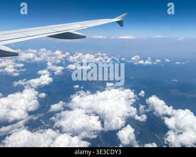 Vue aérienne depuis une fenêtre d'avion montrant une aile de jet au-dessus d'une couche de nuages blancs et moelleux et paysage de montagne en dessous. Banque D'Images