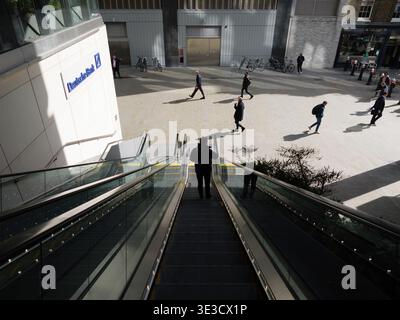 Un navetteur descend les escaliers mécaniques extérieurs à côté du siège de la Deutsche Bank UK au 21 Moorfields. Conçu par WilkinsonEyre et achevé en 2023, ce développement de 17 étages « sur-station » sert de maison unifiée à Londres pour les divisions d'investissement, d'entreprise et de gestion de patrimoine de la banque. Le bâtiment est un exploit d'ingénierie, suspendu au-dessus de l'échangeur de la ligne Elizabeth et de la ligne Northern en utilisant une structure de pont en acier massive. Au premier plan, les piétons marchent le long des Moorfields et de Moor Lane récemment piétons, en admirant le soleil d'hiver qui se reflète sur le bâtiment. » Banque D'Images