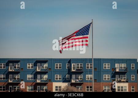 Le drapeau américain flotte sur un mât de drapeau argenté devant un immeuble moderne bleu et brique sous un ciel clair à Wheaton Illinois Banque D'Images