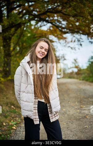 Jeune femme souriante en veste légère debout sur la route forestière d'automne, lumière naturelle chaude, portrait de style de vie décontracté avec fond de feuillage coloré Banque D'Images