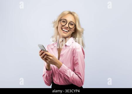 Jeune femme sourit tout en tenant smartphone et en portant des lunettes en arrière-plan clair Banque D'Images
