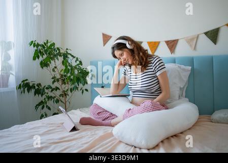 Femme enceinte assistant à un cours prénatal en ligne à la maison, écoutant une conférence sur l'accouchement avec des écouteurs et prenant des notes sur un ordinateur portable tout en étant confortablement assis sur un lit avec un oreiller de grossesse Banque D'Images