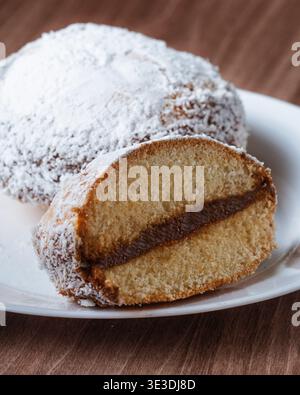 Gâteau à la noix de coco saupoudré de sucre en poudre repose sur une assiette blanche. Slice révèle une garniture riche et chocolatée à l'intérieur d'un gâteau doux et doré. Chaleureux, invitant et déca Banque D'Images