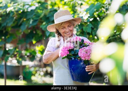 Femme âgée souriante tenant pot de fleurs roses dans le jardin. Banque D'Images