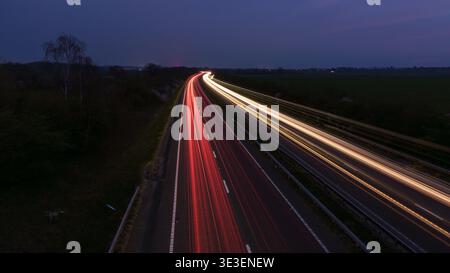 Vue longue exposition de l'autoroute au crépuscule avec des traînées de lumière rouge et blanche de voitures en mouvement, courbant dans la distance à travers la campagne sombre sous TW nuageux Banque D'Images