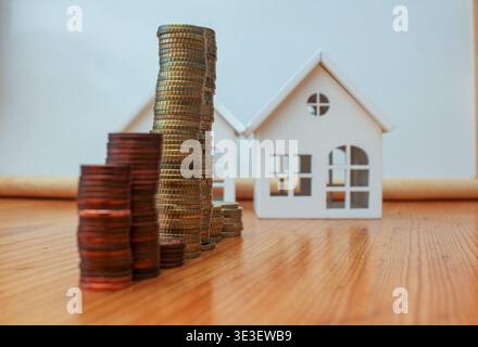 Une image conceptuelle représentant une maison miniature en bois en arrière-plan avec plusieurs piles de pièces de cuivre centimes d'euro au premier plan flou. Ceci i Banque D'Images