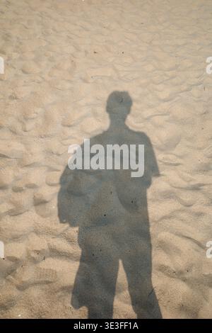 Silhouette d'une personne se dresse sur la plage de sable. Ombre projetée par la figure sur la surface texturée de sable. La lumière du soleil crée un contour distinct et sombre. Personne apparaît Banque D'Images
