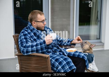 L'homme en robe à carreaux est assis sur le patio avec du café et nourrit son bouledogue français. Routine matinale tranquille à la maison, moment de style de vie confortable avec animal de compagnie. Banque D'Images