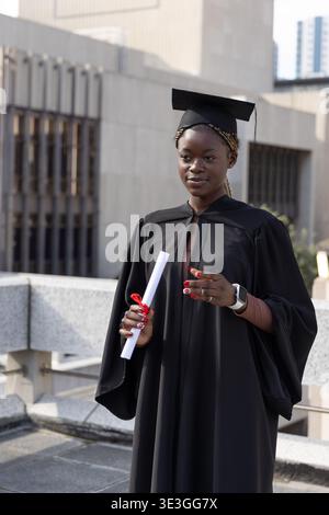 Femme afro-américaine portant robe et casquette debout sur le toit tenant le diplôme roulé avec ruban Banque D'Images