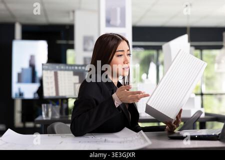 Une professionnelle féminine concentrée tient un modèle architectural blanc tout en discutant avec le client lors d'un appel vidéo. Jeune architecte asiatique évaluant les détails du projet avec une équipe à distance sur le chantier de construction. Banque D'Images