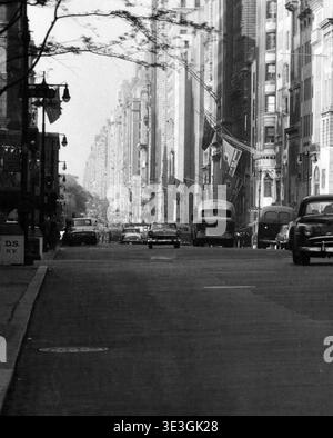 Cette photographie vintage en noir et blanc, probablement capturée par Angelo Rizzuto dans les années 1950, représente une scène de rue animée à New York. Voitures, bus et taxi naviguent sur la large artère urbaine, flanquée de grands bâtiments de la ville et de lampadaires. Banque D'Images