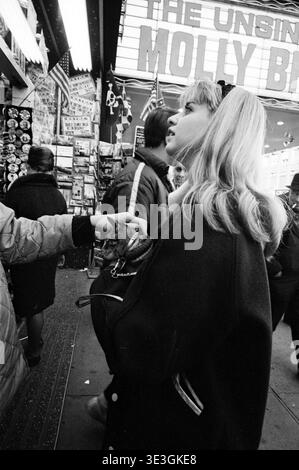 Capturée par Angelo Rizzuto, cette photographie de rue des années 1960 montre une femme blonde tenant un chat dans un porte-chat alors qu'elle passait devant une devanture de New York. La marquise de « l'insubmersible Molly B » (faisant probablement référence à « Molly Brown ») est visible à l'arrière-plan, au milieu de diverses affiches et drapeaux, capturant un moment franc de la vie urbaine. Banque D'Images