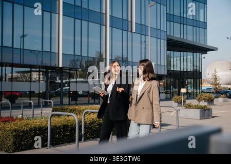 Deux femmes sont engagées dans la conversation alors qu'elles marchent à l'extérieur d'un immeuble de bureaux en verre. Ils portent des vêtements professionnels et semblent concentrés sur leur travail w Banque D'Images