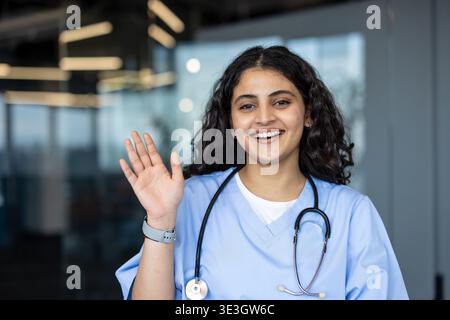 Jeune professionnelle médicale féminine dans les gommages et stéthoscope souriant largement et agitant une main accueillante, représentant les soins de santé, le bien-être et le service amical aux patients Banque D'Images