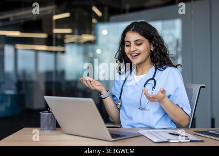 Jeune femme indienne médecin dans les gommages et stéthoscope souriant et gestuel tout en ayant un appel vidéo sur son ordinateur portable, fournissant une consultation médicale en ligne à partir d'un bureau moderne Banque D'Images