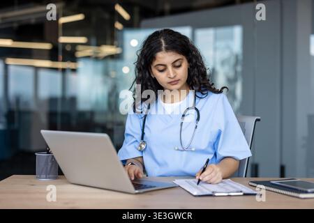 Jeune femme médecin ou infirmière en gommage bleu à l'aide d'un ordinateur portable et écrivant des notes sur un presse-papiers à un bureau, en se concentrant sur la recherche en soins de santé et les tâches de saisie de données Banque D'Images
