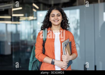 Jeune étudiante indienne portant un sac à dos et des cahiers, debout dans une université moderne ou un immeuble de bureaux, regardant la caméra avec une expression confiante et heureuse Banque D'Images