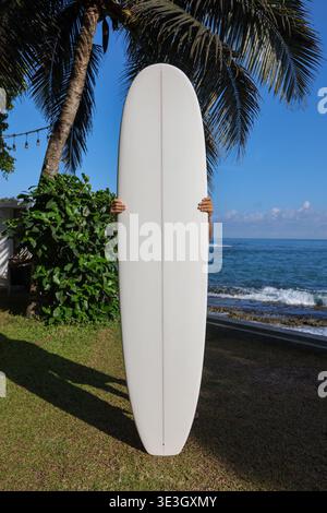 Homme tenant une planche de surf blanche sur une plage tropicale avec vue sur l'océan Banque D'Images