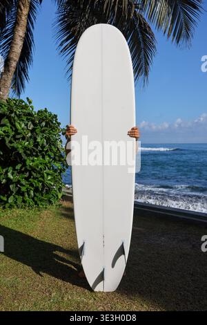 Homme tenant une planche de surf blanche sur une plage tropicale avec vue sur l'océan Banque D'Images