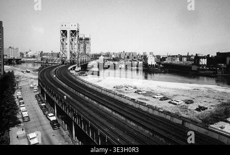 Cette photographie vintage en noir et blanc, probablement des années 1950 par Angelo Rizzuto, capture une vue panoramique de voies ferrées courbées le long d'une rivière, peut-être la rivière Harlem. De nombreuses voitures sont garées le long de la rue et sur un terrain de terre, offrant un aperçu de la vie quotidienne et de la photographie de rue du vieux New York. Banque D'Images