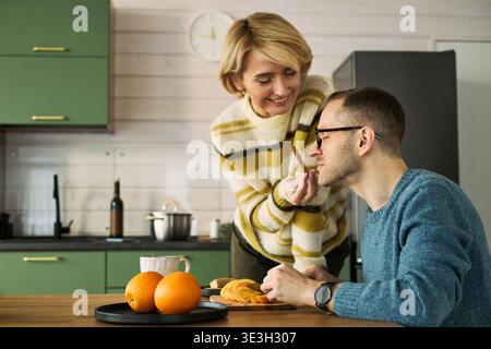 Femme caucasienne d'âge moyen souriant tout en nourrissant la tranche d'orange à l'homme caucasien d'âge moyen assis à la table de la cuisine, les deux interagissant chaleureusement, les oranges et la plaque visibles au premier plan Banque D'Images