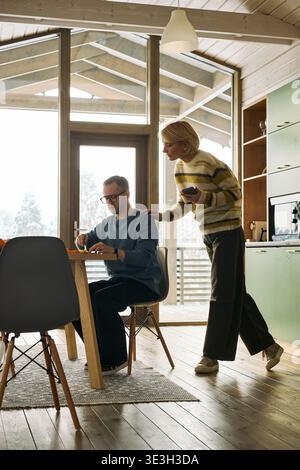 Homme caucasien d'âge moyen assis à table tandis qu'une femme caucasienne d'âge moyen debout à côté de lui, touchant son épaule dans une cuisine moderne en bois, verticale Banque D'Images