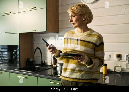 Femme caucasienne d'âge moyen debout dans la cuisine tenant un smartphone, regardant loin de la caméra avec une expression réfléchie Banque D'Images