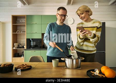 Homme d'âge moyen caucasien remuant le pot sur le comptoir de la cuisine tandis que femme d'âge moyen caucasienne debout à côté de lui tenant un smartphone et souriant à l'écran ensemble Banque D'Images
