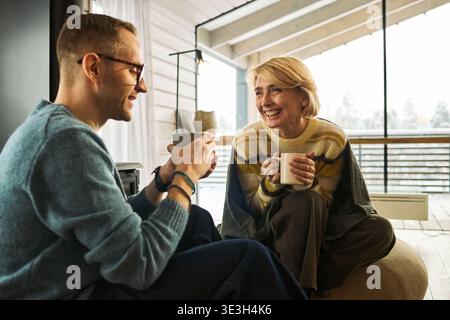 Homme caucasien d'âge moyen et femme caucasienne d'âge moyen assis à l'intérieur souriant et tenant des tasses, s'engageant dans une conversation amicale près d'une grande fenêtre avec la lumière naturelle qui coule dedans Banque D'Images