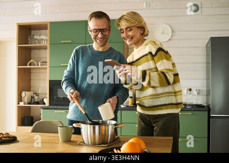 Homme d'âge moyen caucasien remuant le pot sur la cuisinière tout en souriant femme d'âge moyen caucasien tenant un smartphone et montrant l'écran, les deux debout ensemble dans un cadre de cuisine moderne Banque D'Images
