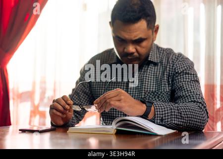 Un homme d'âge moyen dans une chemise à carreaux tenant ses lunettes tout en se concentrant sur la lecture d'un livre, dépeignant la concentration, l'étude et la sagesse. Banque D'Images