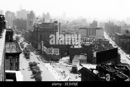 Ce magnifique paysage urbain, capturé par le photographe Angelo Rizzuto au milieu des années 1950, présente une vue aérienne d'une rue animée de New York. La photographie en noir et blanc met en valeur la densité architecturale de la ville avec de nombreux bâtiments, des voitures anciennes et l'emblématique Empire State Building subtilement visible dans la distance brumeuse. Banque D'Images