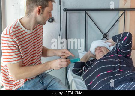 Le fils adulte donne des médicaments à la mère malade se reposant dans le lit, vérifiant son état et soutenant le rétablissement. Soins familiaux pour les parents âgés, soutien et Banque D'Images