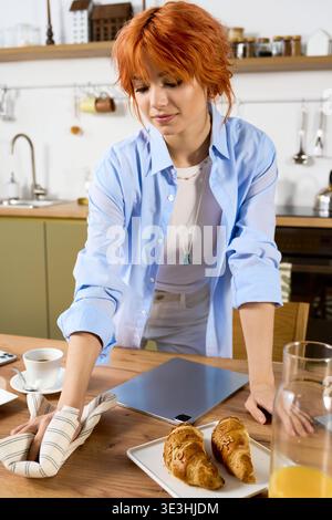 Femme aux cheveux rouges aime une routine matinale, essuyant la table tout en préparant des pâtisseries fraîches à la maison. Banque D'Images