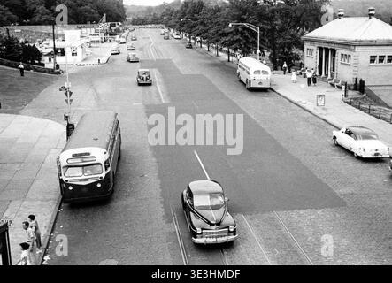 Cette scène de rue en noir et blanc, capturée par le photographe Angelo Rizzuto au milieu des années 1950, dépeint la vie quotidienne à New York avec de nombreux véhicules anciens, dont des bus et des voitures, traversant une large route. Les piétons sont visibles sur les trottoirs, et un bâtiment classique en pierre avec des colonnes se dresse sur la droite, mettant en valeur la mode et l'environnement urbain de l'époque. Banque D'Images