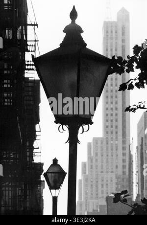 Cette superbe photographie en noir et blanc d'Angelo Rizzuto, probablement des années 1940 ou 1950, capture une vue détaillée d'un lampadaire classique de New York au premier plan, silhouette contre un haut gratte-ciel. La composition souligne le contraste entre le mobilier urbain ancien monde et le paysage urbain moderne, incarnant l'essence intemporelle de la photographie de rue New-yorkaise vintage. Banque D'Images