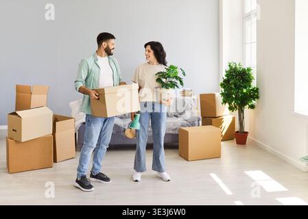 Couple heureux emménageant dans une nouvelle maison, transportant des boîtes et des plantes d'intérieur ensemble Banque D'Images