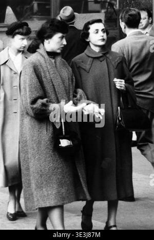 Cette photographie de rue vintage, probablement capturée par Angelo Rizzuto dans les années 1950, représente deux femmes bien habillées marchant dans une rue animée de New York, mettant en valeur la mode et la vie quotidienne de l'époque. Leurs manteaux et accessoires élégants reflètent l'élégance urbaine du milieu du siècle de Manhattan. Banque D'Images