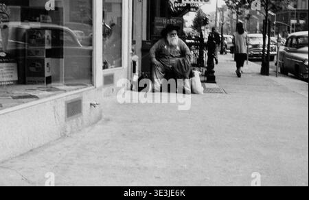 Photographie de rue en noir et blanc des années 1950 par Angelo Rizzuto, montrant un homme âgé barbu assis sur un trottoir de New York. L'homme, peut-être un commerçant ou un résident, est positionné devant une vitrine de magasin avec des reflets de rue, offrant un aperçu de la vie quotidienne et de la mode d'époque de New York vintage. Banque D'Images