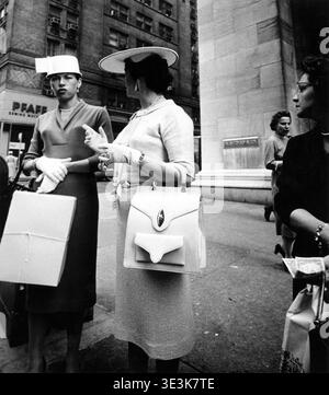 Deux femmes bien habillées, photographiées par le photographe Angelo Rizzuto, sont engagées dans une conversation dans une rue de New York dans les années 1950 Ils illustrent la mode élégante et la vie de rue de l'époque, présentant des chapeaux élégants, des gants et des sacs à main tout en étant entourés par l'architecture urbaine. Banque D'Images