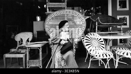 Des années 1950, cette photographie de rue réalisée par Angelo Rizzuto capture une femme âgée assise confortablement dans une grande chaise paon en osier devant un magasin de meubles à New York. Elle tient un journal, son sac à main et une canne, incarnant un moment de répit et de vie quotidienne dans une rue de ville. Banque D'Images