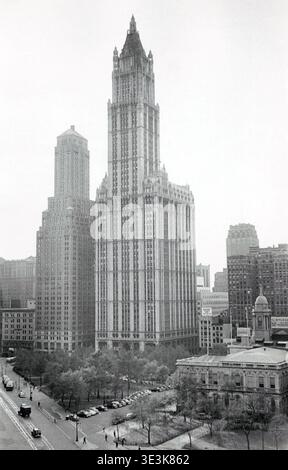Une vue aérienne impressionnante capture l'emblématique Woolworth Building surplombant une scène de rue animée à New York, probablement des années 1950, photographiée par Angelo Rizzuto. Les voitures anciennes bordent les rues et les gens se promènent dans un parc au premier plan, mettant en valeur la vie quotidienne et l'architecture urbaine du milieu du XXe siècle. Banque D'Images